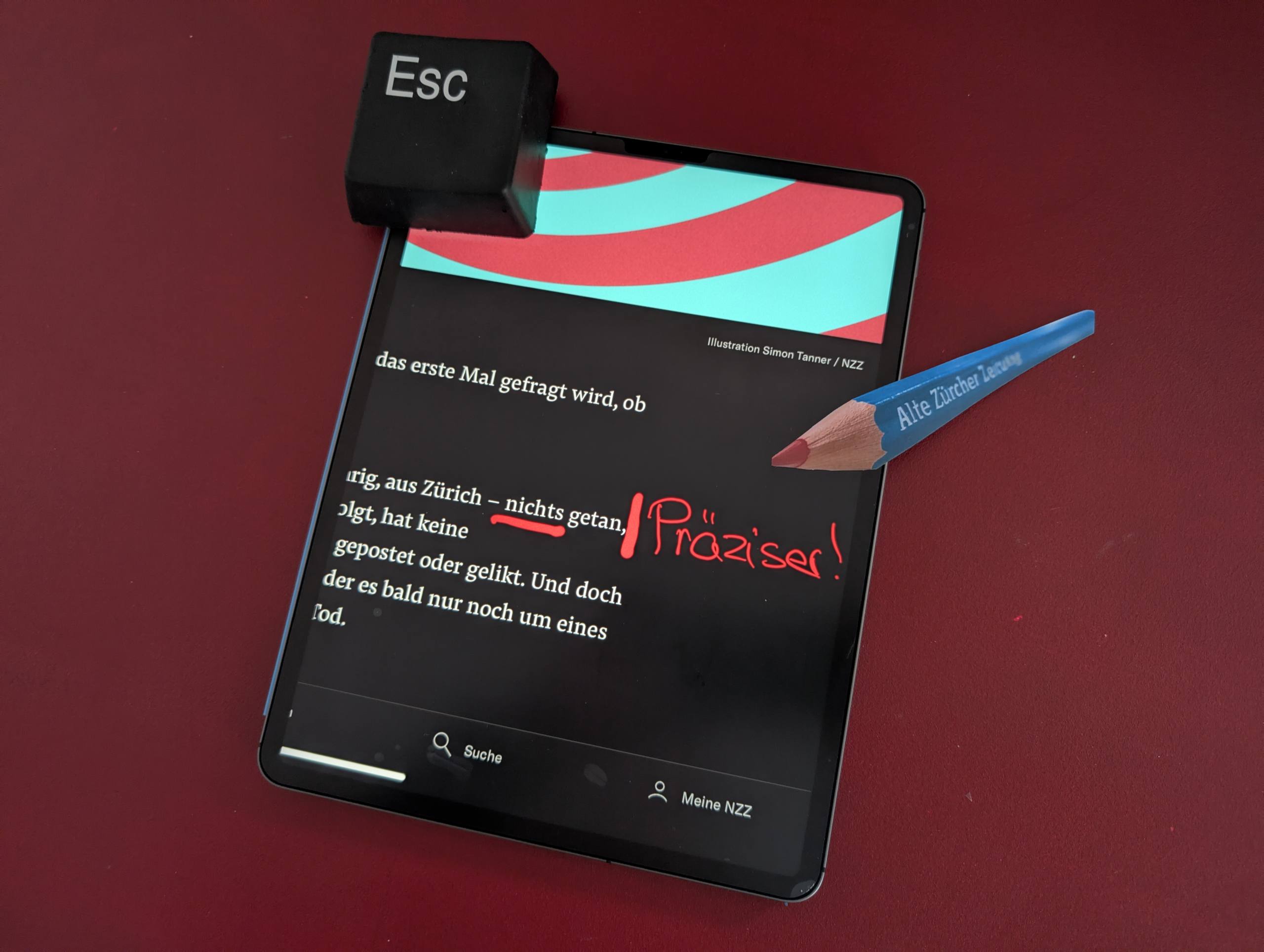 Auf dem Bild ist ein Tablet zu sehen, das auf einer roten Oberfläche liegt. Auf dem Bildschirm des Tablets ist ein Text in deutscher Sprache zu lesen, der folgendermassen lautet: "In diesen zehn Minuten hat Lena – 14-jährig, aus Zürich – nichts getan, ausser zu scrollen." Das Wort "nichts" ist von Hand rot unterstrichen. Daneben steht von Hand geschrieben das Wort "Präziser!" Neben dem Tablet liegt ein grosser blauer Bleistift mit roter Mine, auf dem die Aufschrift "Alte Zürcher Zeitung" in weißer Schrift zu sehen ist. Oben links auf dem Tablet liegt ein schwarzer Würfel mit der Aufschrift "Esc". Das ist eine grosse Spielzeug-Escape-Taste.