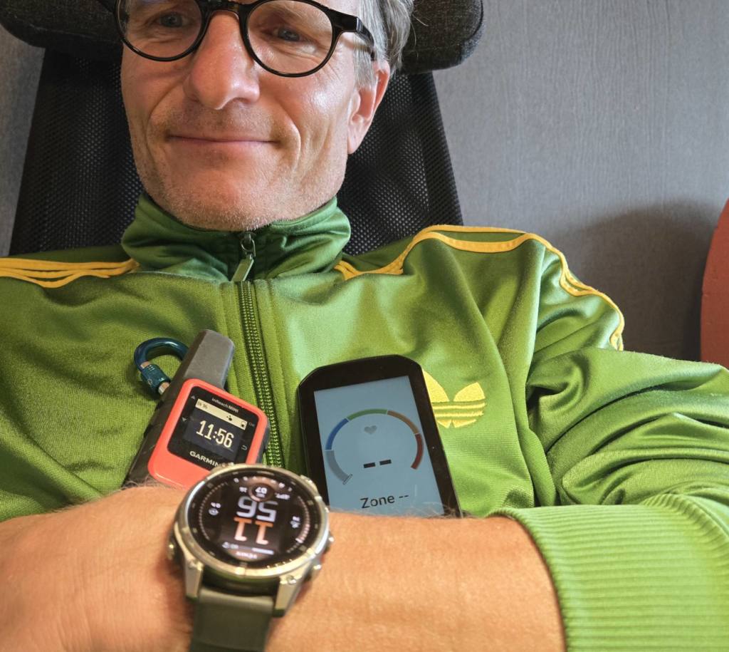 Marc Böhler mit Garmin Tabs: Eine Garmin Fenix Smartwatch (Gesundheits- und Aktivitätstracking), ein Garmin InReach Mini (Satelliten-Notfallkommunikation) und ein Garmin Edge Velocomputer (Leistungsdaten für Radfahrer). Die Geräte symbolisieren vernetzte, alltagsintegrierte Technologie für Gesundheit, Sicherheit und soziale Verbindung.