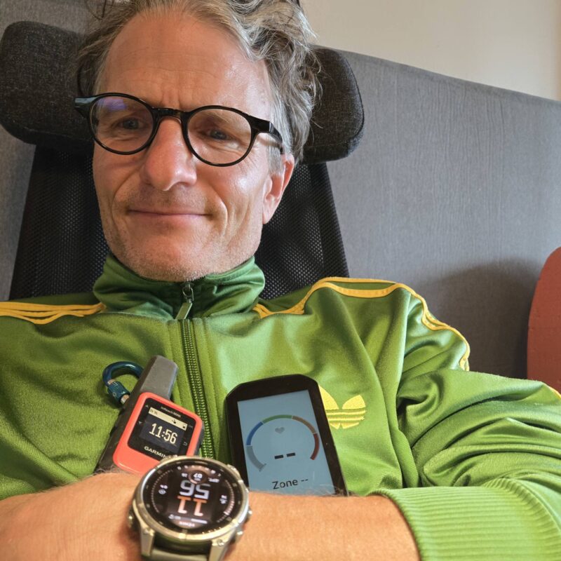 Marc Böhler mit Garmin Tabs: Eine Garmin Fenix Smartwatch (Gesundheits- und Aktivitätstracking), ein Garmin InReach Mini (Satelliten-Notfallkommunikation) und ein Garmin Edge Velocomputer (Leistungsdaten für Radfahrer). Die Geräte symbolisieren vernetzte, alltagsintegrierte Technologie für Gesundheit, Sicherheit und soziale Verbindung.